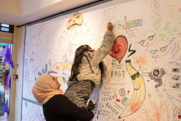 A mother holds her young daughter in front of her as she reaches to draw on a giant sheet of paper on the wall