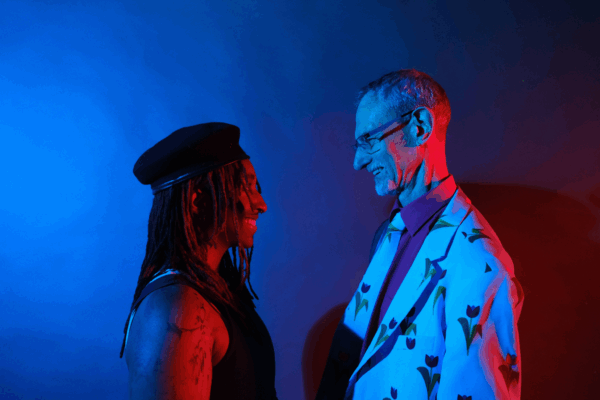 Blue lit film photo - a young black person with dreads and a hat looks up to a white man wearing a suit covered in cartoon tulips.