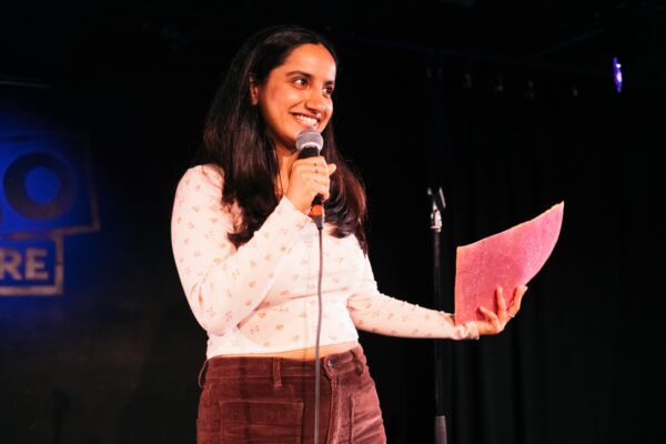 An Indian woman with long dark hair poses on stage - she is holding a microphone and some pink folders, and is wearing brown trousers and a white top.