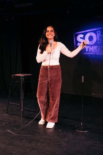 An Indian woman with long dark hair poses on stage - she is holding a microphone and is wearing brown trousers and a white top.