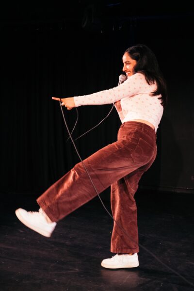 An Indian woman with long dark hair poses on stage - she is holding a microphone and is wearing brown trousers and a white top.