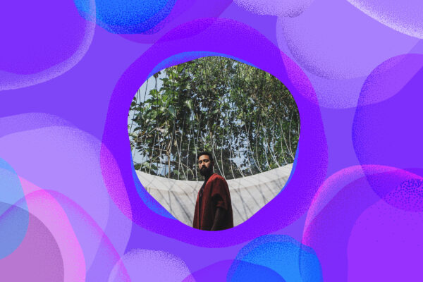 A man wearing flowing red clothes looks directly towards us. Behind him there are trees in leaf and a blue sky.