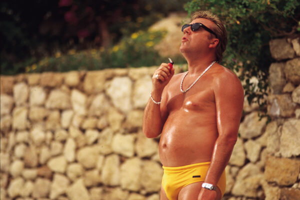 A man standing in yellow speedos smokes a cigarette.