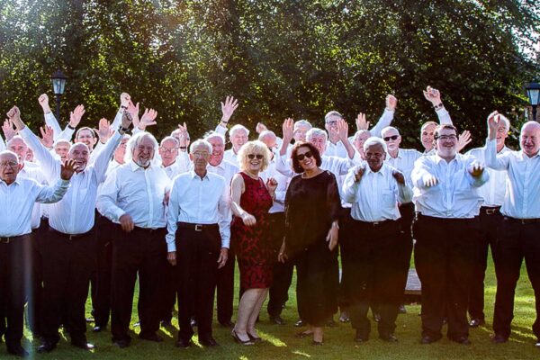 A large group of men in black trousers and white shirts cheer in a park. Two women are stood at the front in dresses.
