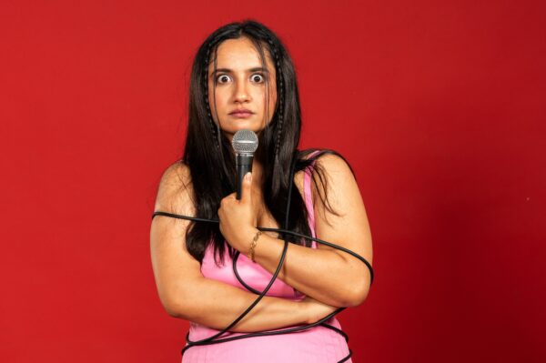 A woman with long dark hair and wearing a pink dress stares into the camera - her background is red and she's holding a microphone.