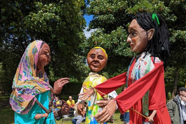 Three giant puppets of women from different cultural backgrounds perform in a park