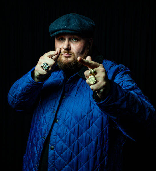 Rapper, Big Deli, wearing a flat cap pointing towards the camera. Wearing a blue jacket.