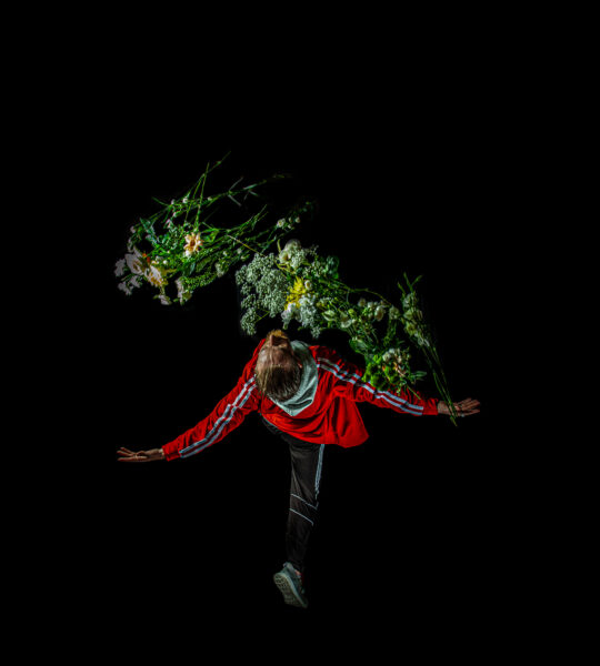 Male in red sports jacket leaning back, with a branch that has leaves and flowers on.