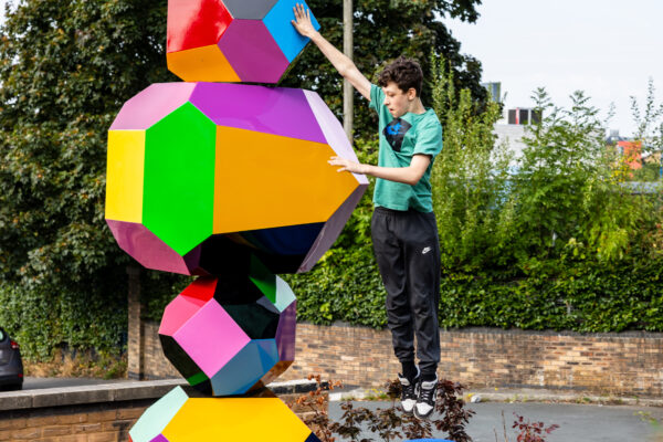 Young person in mid air next to the colourful obelisk-shaped structure, Balancing Acts
