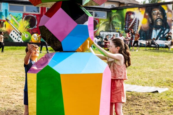 Two young people touching the bottom shape of the colourful obelisk-shaped structure