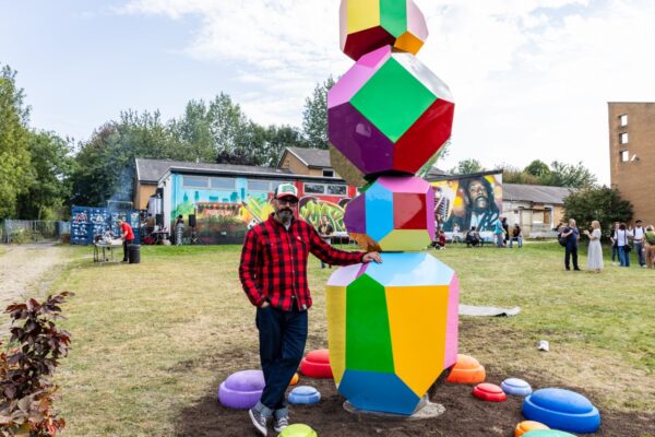 Artist, Sam Shendi standing next to Balancing Acts. A colourful obelisk-shaped structure