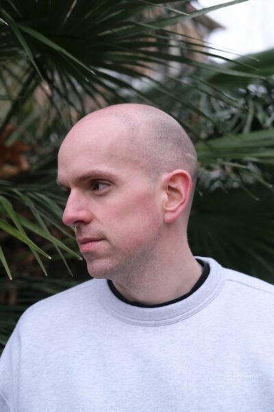 A white man poses for the camera - he is wearing a pale grey sweater and the background is green plants.