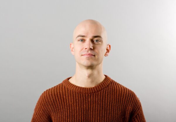 A white man poses for the camera - he is wearing a red jumper and the wall behind him is white.