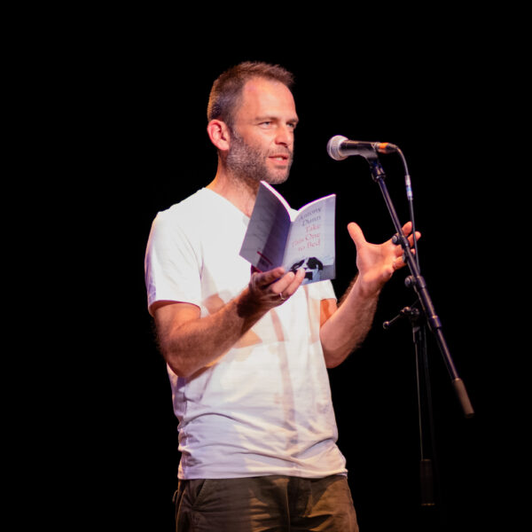 A white man reads from a book into a microphone - he is wearing a white t-shirt and the background is black.