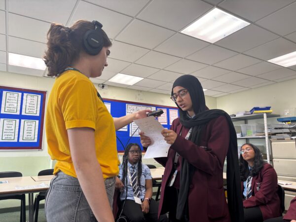 One person, wearing a yellow t-shirt, holds up the microphone to a school girl as she reads from a sheet of paper.