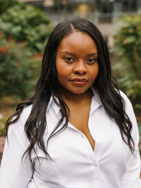 A black woman with long hair poses for the camera, she is wearing a white shirt and there are plants in the background