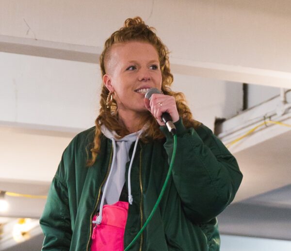 Poet Kirsty Taylor wearing a green jacket performs using a microphone.