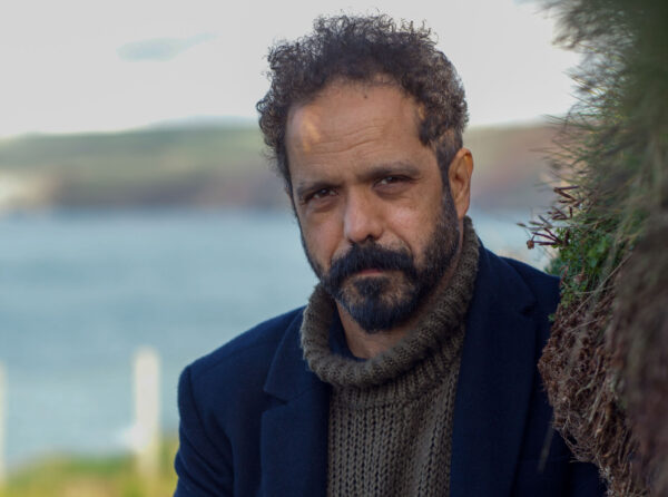 A man with curly hair and a beard looks into the camera - he is wearing a dark jacket and a grey jumper, the background is a landscape with a body of water.