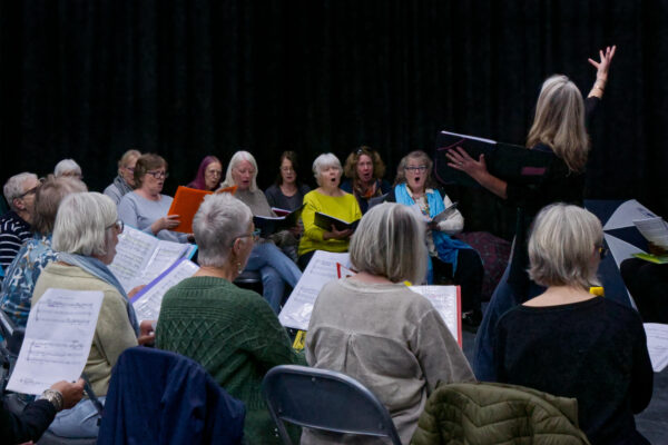 A choir of people practice - conducted by a woman, her hand in the air