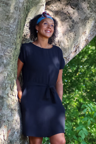 A woman poses for the camera - she has curly hair and is wearing a blue dress as she leans on a tree.