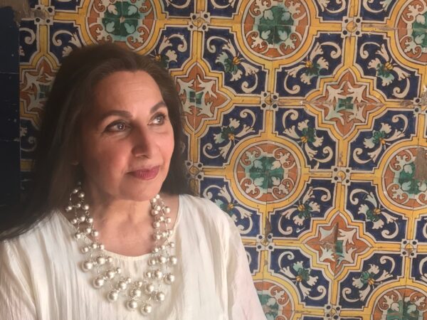 A woman poses in front of a patterned wall - she has dark hair and is wearing a white shirt with a layered necklace.