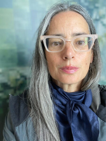 A white woman with grey hair and glasses poses for the camera - she is wearing a blue top and the background is soft blues.