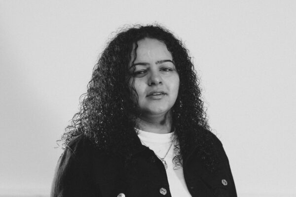 A black and white photo - a woman with long curly hair poses for the camera, she is wearing a pale t-shirt and a dark jacket.