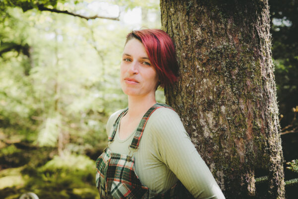 A white woman with red hair poses for the camera - she is wearing a green t-shirt and a plaid top, and is leaning against a tree.
