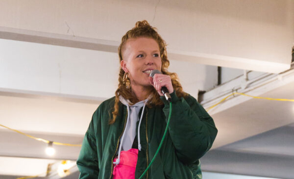 Poet Kirsty Taylor wearing a green jacket performs using a microphone.