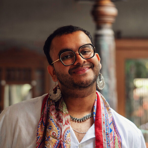 A person poses for the camera - they have round glasses on, intricate earrings and a colourful scarf over their shirt