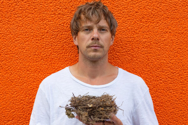 A man poses for the camera - he has brown hair, is wearing a white t shirt and the background is orange