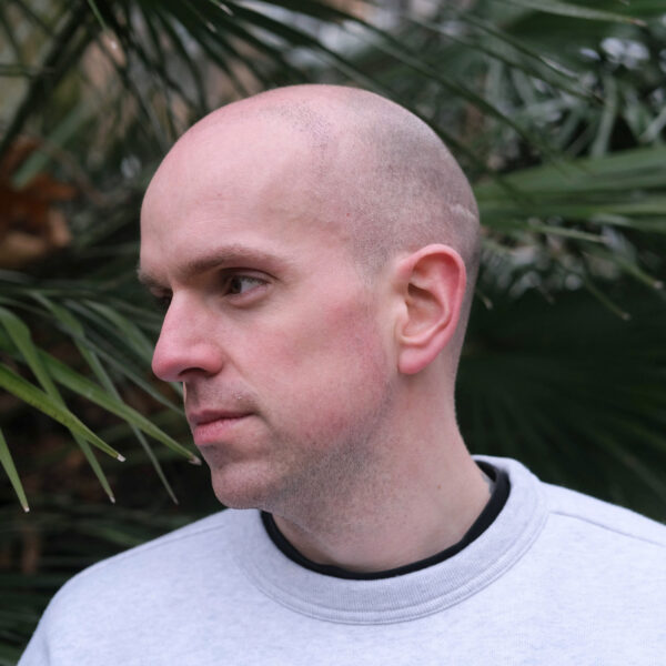 A headshot of a man - he is wearing a grey sweater and is looking to one side, posing in front of plants.