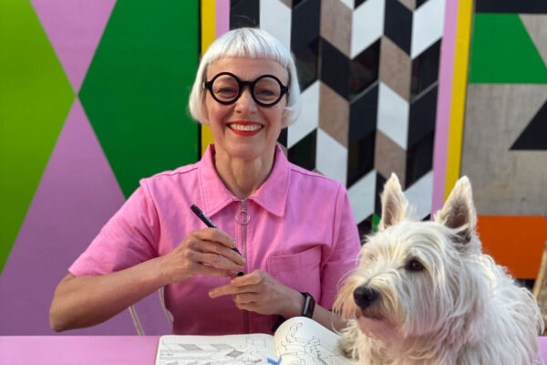 Artist Morag Myserscough and her dog Elvis. Morag has a silver bob and round black glasses and wears a pink top. She is sitting at a desk with a notebook. Elvis is a very handsome white terrior dog.