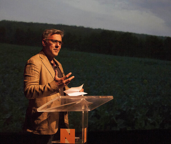 A man reads at a podium - he is wearing a suit and there is a screen behind him.