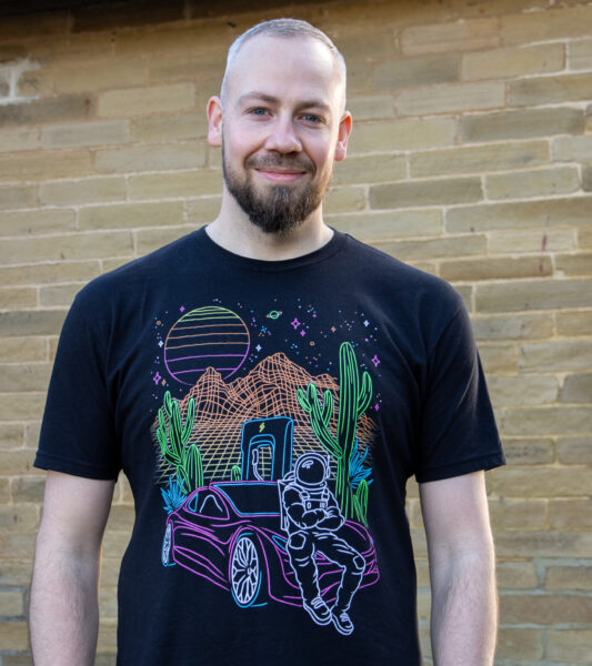A white man poses for the camera - he is smiling, wearing a t-shirt and there is a brick wall behind him.