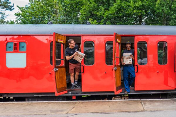 Two people looking out of the doors of a red train carriage, holding newspapers.