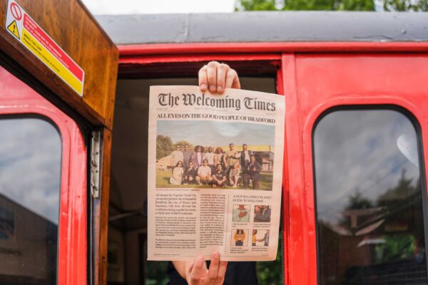 A person holding out a newspaper through a red train door. The newspaper headline reads 'The Welcoming Times'.