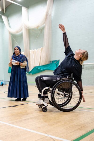 Three performers in a rehearsal. A dancer on the left kneeling. A woman speaking in the centre. And a dancer in a wheelchair on the right.