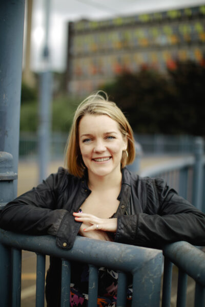 A white woman smiles at the camera as she leans against a set of grey railings - she has blonde hair and is wearing a black jacket.