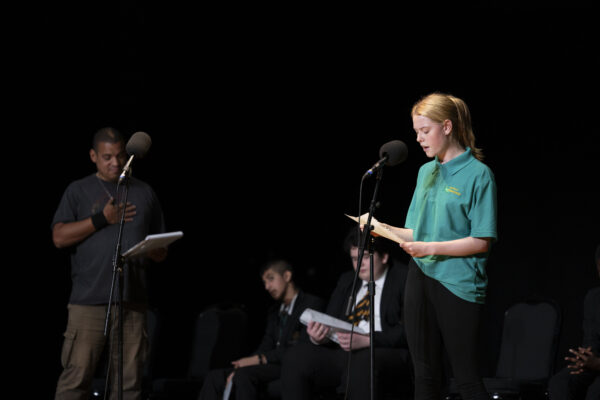 A young girl performs - reading into a microphone, she is wearing a green uniform.
