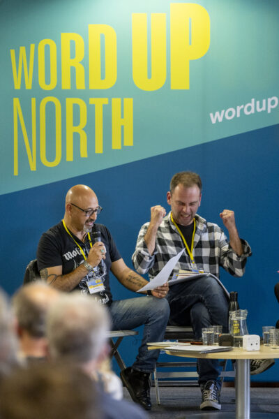 Two men sit on the stage - the one on the left is reading, while the other is cheering.