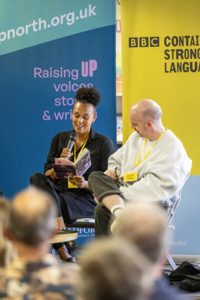Two people sit on the stage - the woman on the left is reading from a book, the man on the right is looking to her.