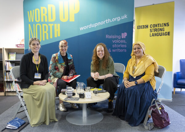 Four women pose for a photo on stage - a big blue physical screen behind them that reads: Word Up North.