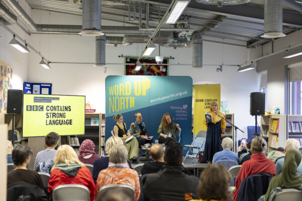 Three women sit on stage as another woman reads into the microphone - a big blue physical screen behind them that reads: Word Up North.