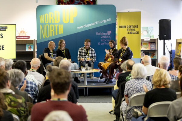Five people sit on the stage before a crowd - a big blue physical screen behind them that reads: Word Up North.