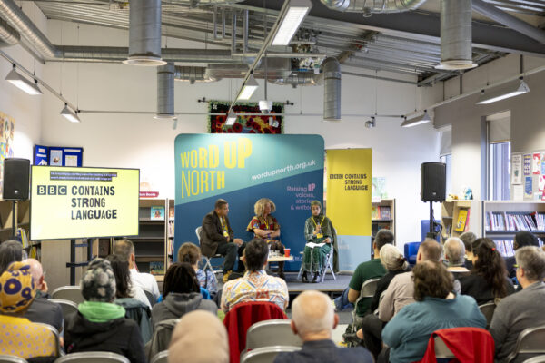 Three people sit on the stage before a crowd - a big blue physical screen behind them that reads: Word Up North.