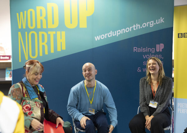 Three people laugh on stage - big blue physical screen behind them that reads: Word Up North.