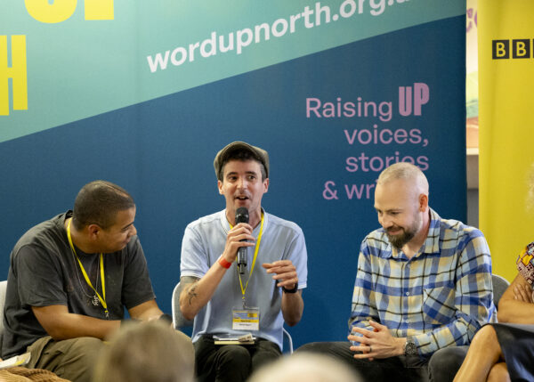A white man talks into the microphone - a big blue physical screen behind them that reads: Word Up North.