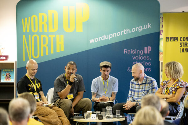 A black man talks into the microphone - a big blue physical screen behind them that reads: Word Up North.