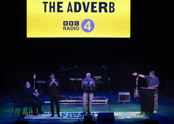 People perform on stage - behind them, a giant yellow screen reads: The Adverb.